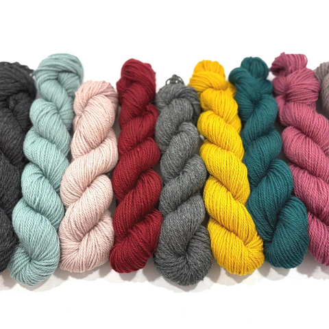 A row of twisted yarn skeins in multiple colors, including black, light blue, blush pink, burgundy red, charcoal gray, bright yellow, teal, and magenta, arranged on a white background.