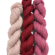 Three twisted skeins of yarn in soft neutral, deep red, and dusty rose tones, arranged vertically on a plain white background.