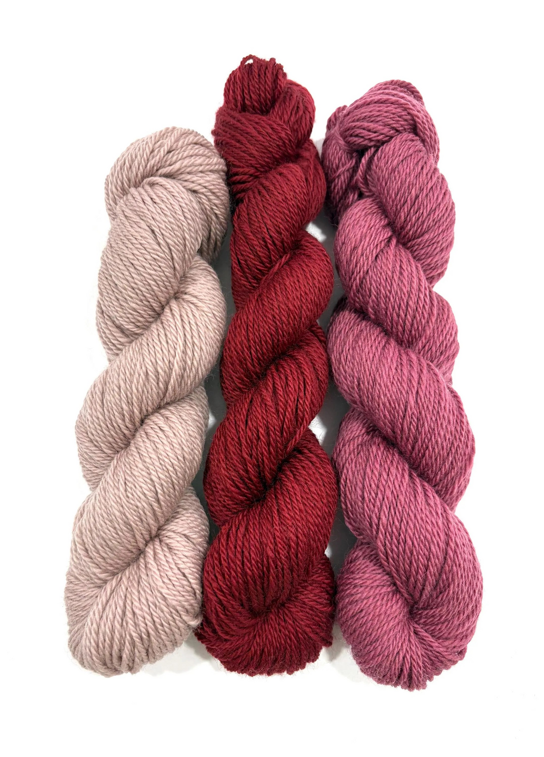 Three twisted skeins of yarn in soft neutral, deep red, and dusty rose tones, arranged vertically on a plain white background.