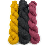 Three skeins of twisted yarn standing vertically on a white background, in mustard yellow, charcoal gray, and dusty rose.