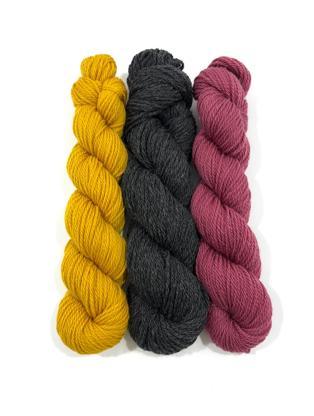 Three skeins of twisted yarn standing vertically on a white background, in mustard yellow, charcoal gray, and dusty rose.