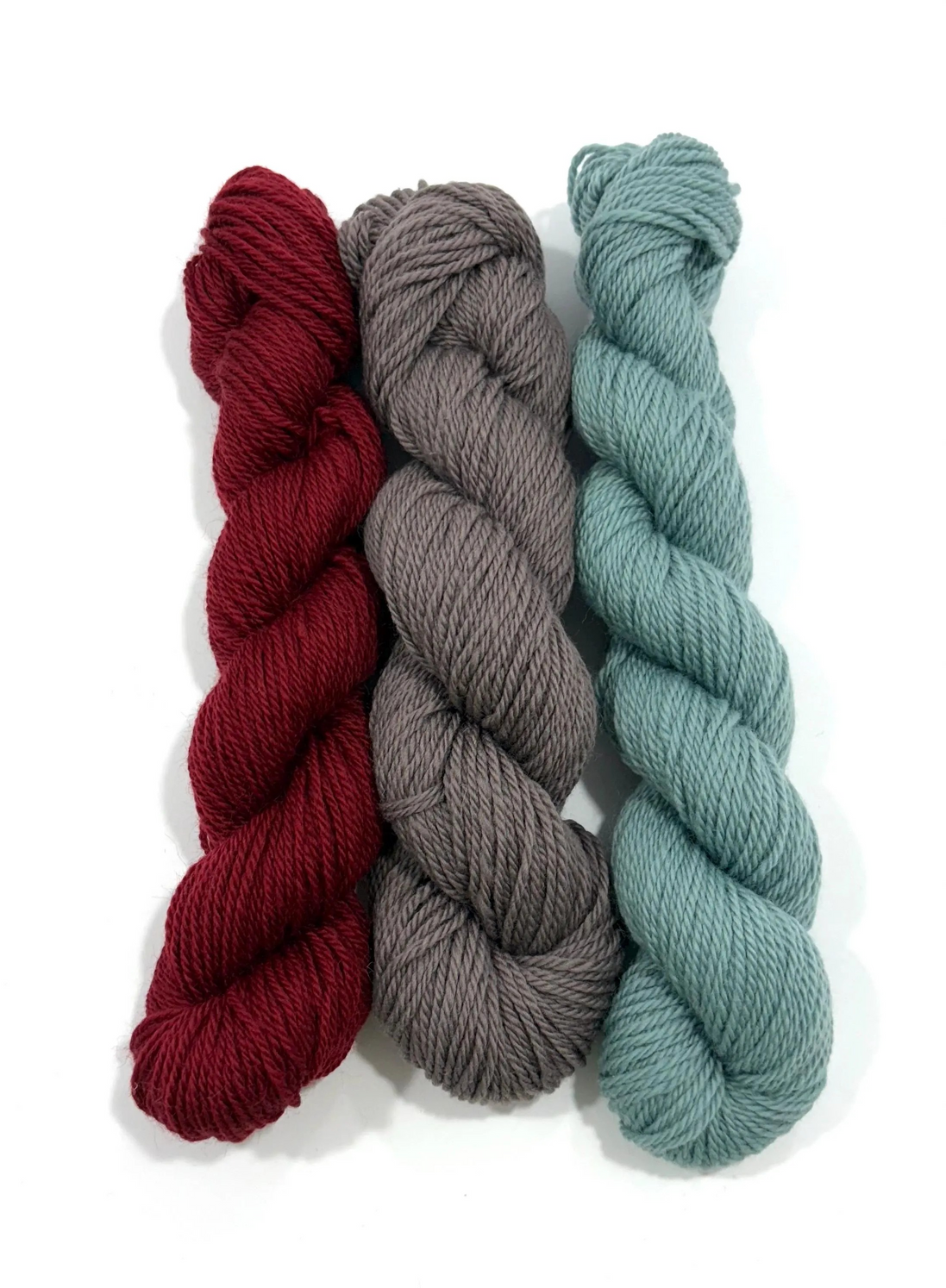 Three skeins of twisted yarn in burgundy red, charcoal gray, and dusty sage green, arranged vertically on a white background.