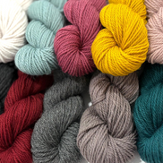 A close-up arrangement of multiple yarn skeins in assorted colors, including cream, pale blue, dusty pink, mustard yellow, blush, charcoal, deep red, taupe, and teal, filling the frame.