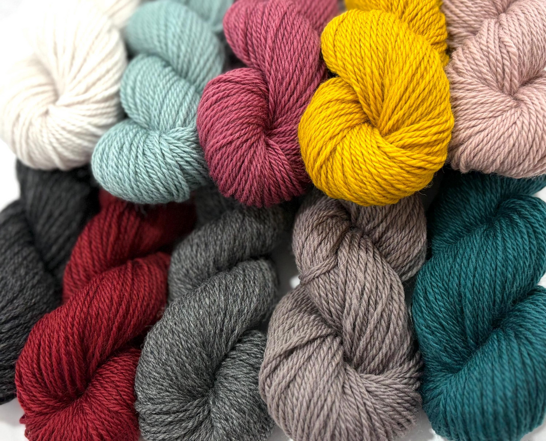 A close-up arrangement of multiple yarn skeins in assorted colors, including cream, pale blue, dusty pink, mustard yellow, blush, charcoal, deep red, taupe, and teal, filling the frame.