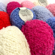 A close-up of several textured boucle or chenille yarn balls in assorted colors, including bright pink, blue, red, cream, and lavender. A paper label on one blue ball reads Gepard Teddy Bear.