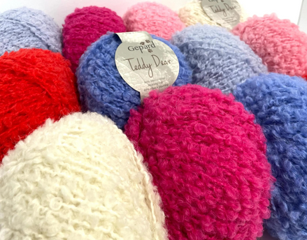 A close-up of several textured boucle or chenille yarn balls in assorted colors, including bright pink, blue, red, cream, and lavender. A paper label on one blue ball reads Gepard Teddy Bear.