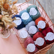 A top-down view of a copper wire basket filled with multiple skeins of yarn in assorted colors, arranged on a round pink table with dried flowers in the corner.