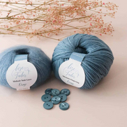 Two balls of dusty blue yarn with white labels reading 'Rose Gold' and 'Midnight Slate Lace', styled with a few matching buttons and dried pink flowers on a light beige background.