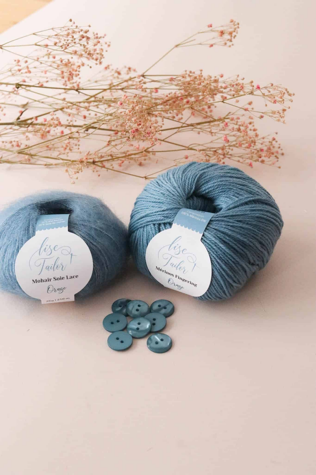 Two balls of dusty blue yarn with white labels reading 'Rose Gold' and 'Midnight Slate Lace', styled with a few matching buttons and dried pink flowers on a light beige background.