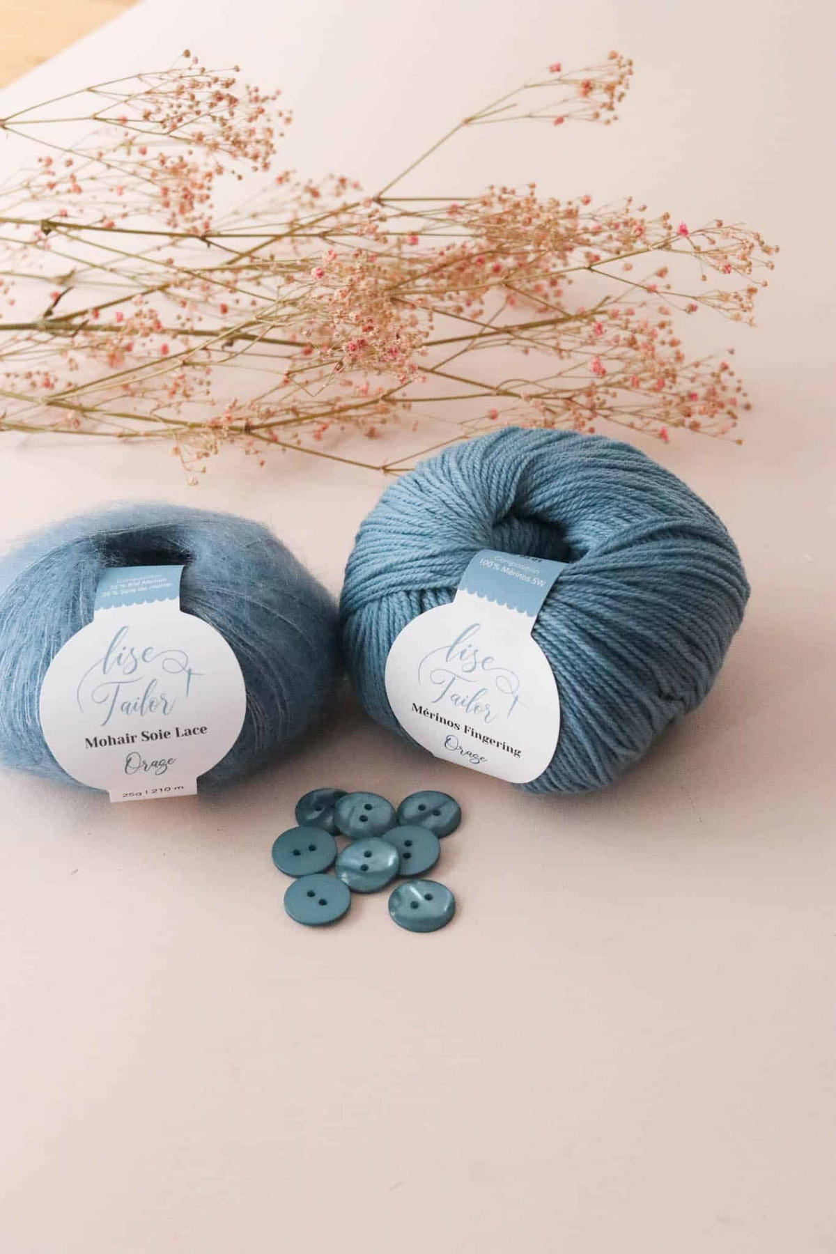 Two balls of dusty blue yarn with white labels reading 'Rose Gold' and 'Midnight Slate Lace', styled with a few matching buttons and dried pink flowers on a light beige background.