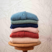 A stack of five folded knit cardigans in light blue, sage green, navy, dusty pink, and terracotta red, displayed on a round wooden stool against a light floral wallpaper background.