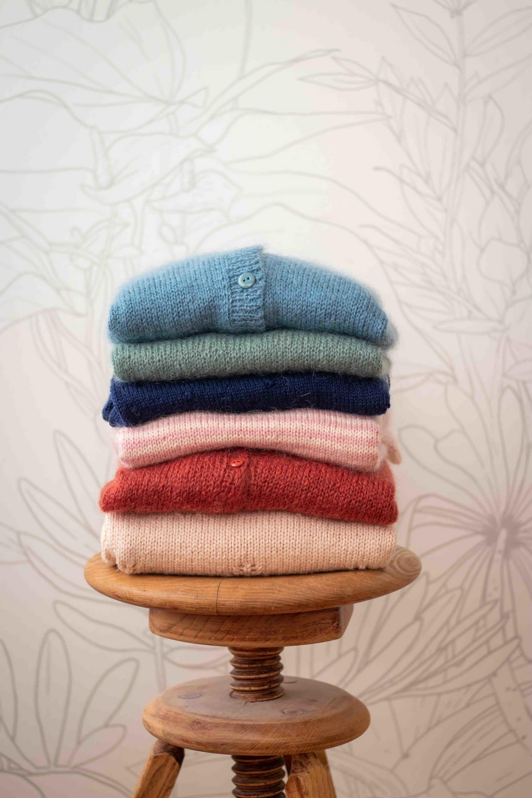 A stack of five folded knit cardigans in light blue, sage green, navy, dusty pink, and terracotta red, displayed on a round wooden stool against a light floral wallpaper background.