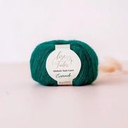 A ball of emerald green mohair sole lace yarn with a white paper label reading 'Lise Tailor' and 'Emerald', photographed on a soft light pink background.