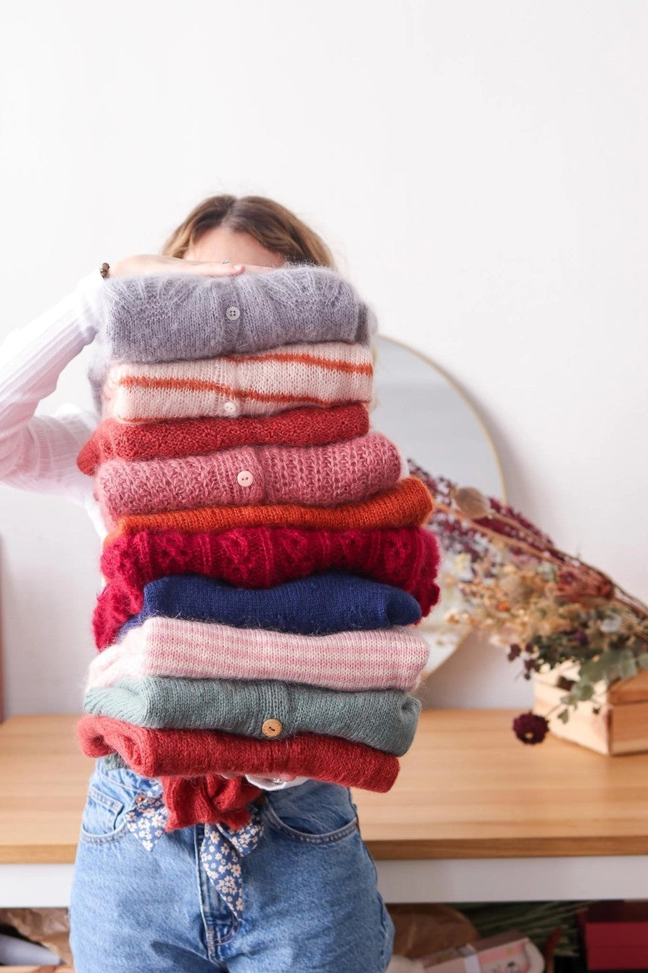 Person holding a stack of colorful sweaters in front of a white wall.
