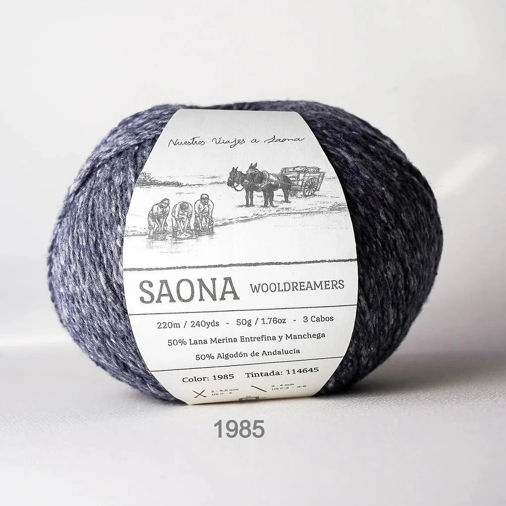 A ball of SAONA Wooldreamers yarn in color 1985, featuring a white paper label with black text and an illustration of people and a donkey. The yarn is a heathered blend of navy and gray-blue tones, photographed on a neutral light gray background.