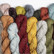 A row of twisted yarn skeins in multiple colors, including black, gray, beige, orange, burgundy, yellow, blue, olive green, and mauve, arranged against a plain white background.