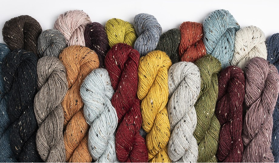 A row of twisted yarn skeins in multiple colors, including black, gray, beige, orange, burgundy, yellow, blue, olive green, and mauve, arranged against a plain white background.
