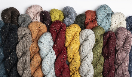 A row of twisted yarn skeins in multiple colors, including black, gray, beige, orange, burgundy, yellow, blue, olive green, and mauve, arranged against a plain white background.